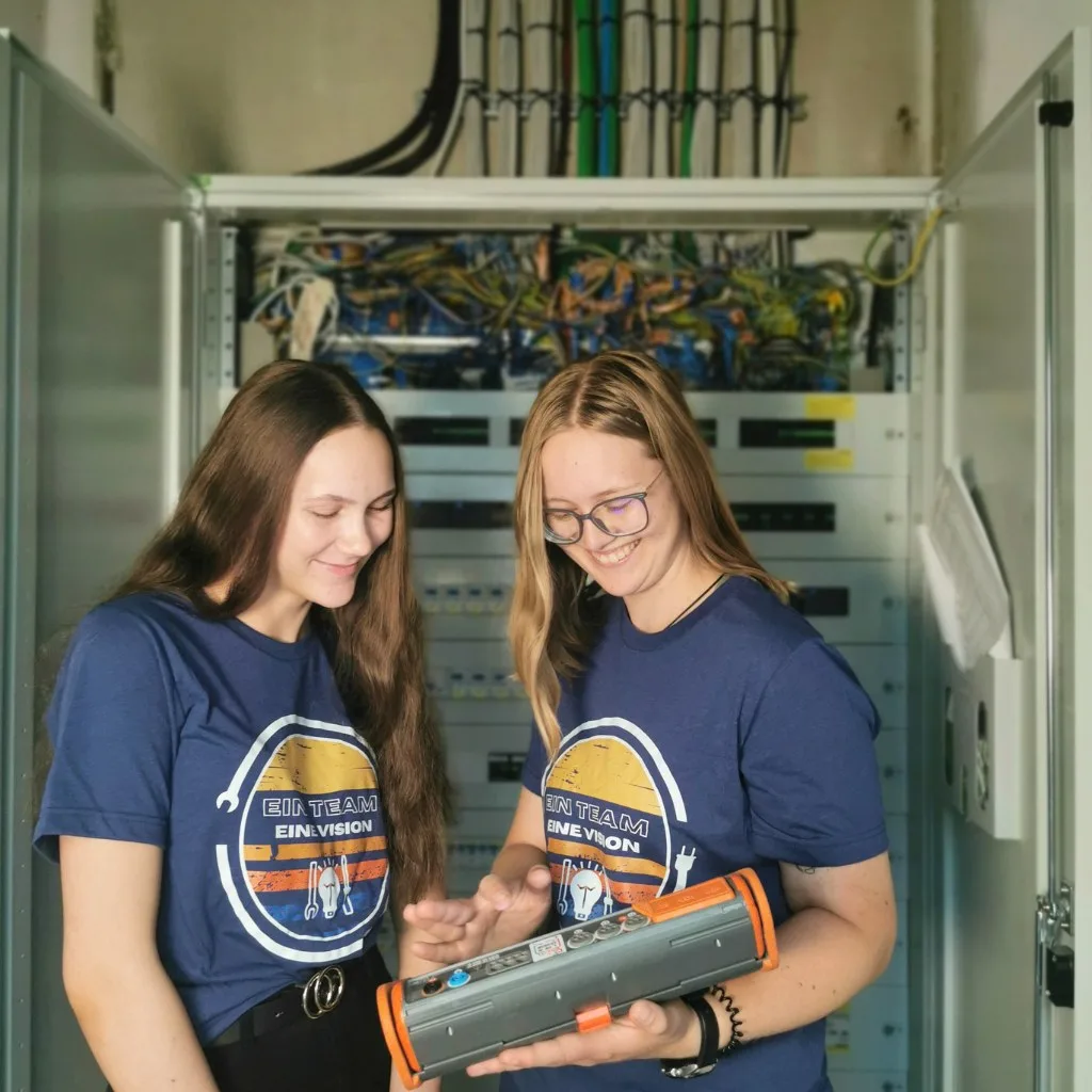 Zwei junge Frauen in Team-Shirts mit einem Messgerät vor einem offenen Schaltschrank